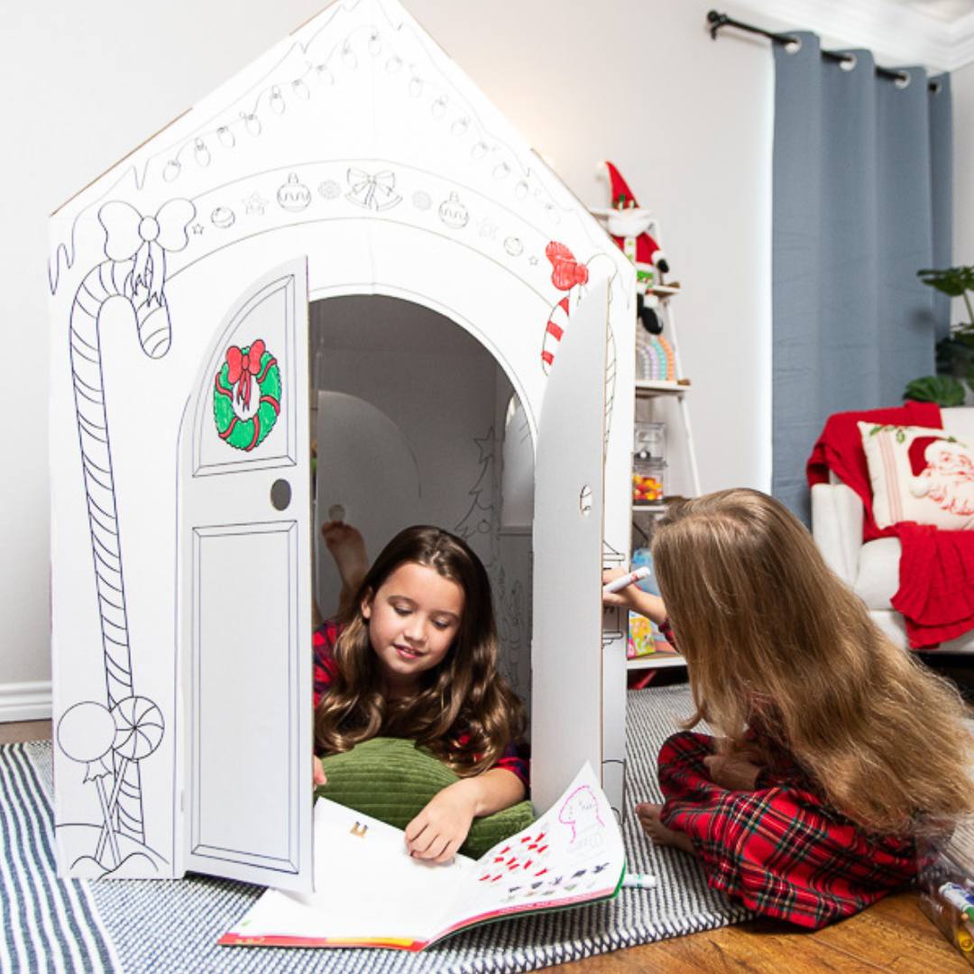 Playboxes Santa’s Workshop playhouse image showing a girl laying inside the Santa’s Workshop Christmas playhouse looking at the activity booklet and a girl sitting outside the Santa’s Workshop Christmas playhouse coloring the outside of the box 